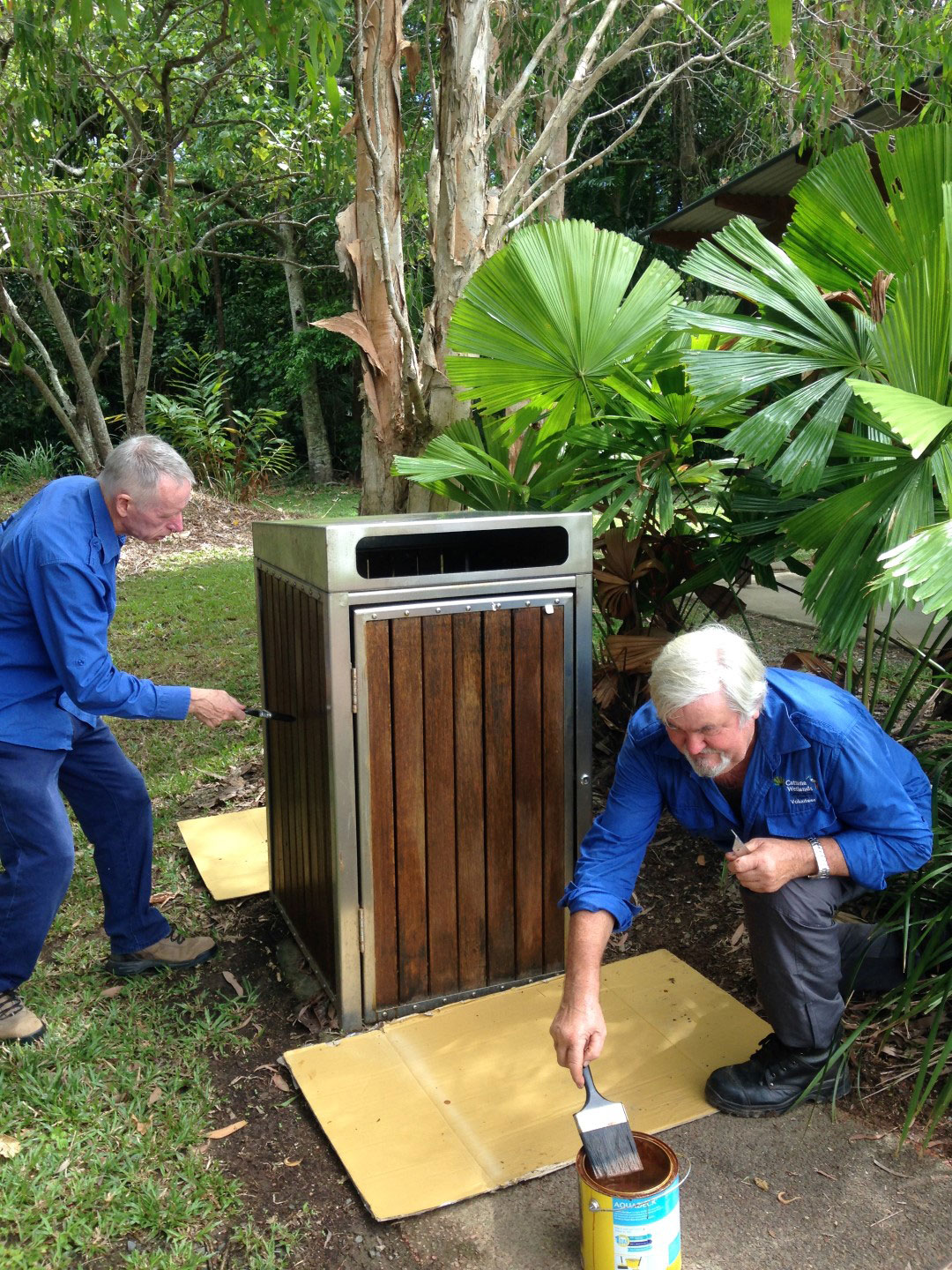 Two volunteers paint new stain on wooden bin surround