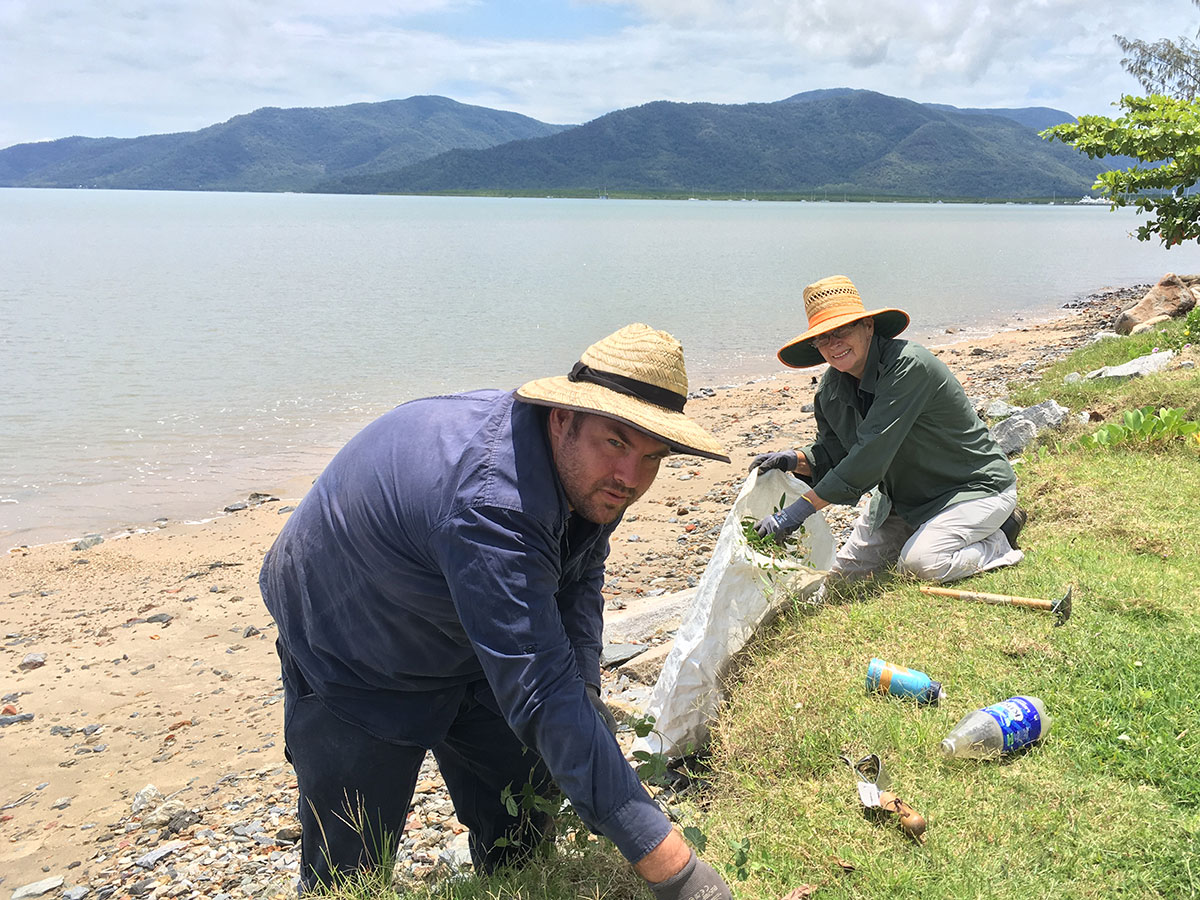 Man in foreground bending down to collect rubbish, woman kneeling in background collecting rubbish on the Cairns Esplanade foreshorre